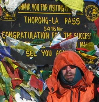 Thomas Münch steht in Bergsteigerklamotten vor einem Glückwunsch-Schild, dass sich auf den Erfolg der Bezwingung des Thorong-La Pass bezieht.
