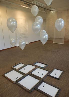 Ein Foto zeigt einen Raum. Im Vordergrund sind weiße, mit Helium gefüllte Luftballons an Fäden an deren unterem Ende schwarze Filzstifte gebunden sind. Sie zeichnen bei jedem Luftstoß auf das darunter liegende Papier.
