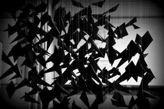 Aus schwarzem Papier gefaltete Vögel an durchsichtigen Fäden. Sie hängen von der Decke herab. Es sind so viele, dass sie einen Schwarm ergeben.
