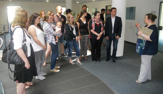 Eine Gruppe Studierender steht halbkreisförmig in einem Abstand von ca. zwei Metern in einem Flurbereich des Jobcenters Düsseldorfs vor einer Mitarbeiterin desselbigen, die einen (kurzen) Vortrag hält.