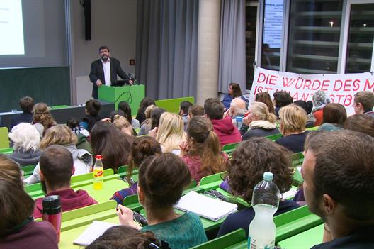 Michael Windfuhr hält einen Vortrag in einem Hörsaal mit grünen Bänken und Tischen und einem Transparent: Die Würde des Menschen ist unantastbar.