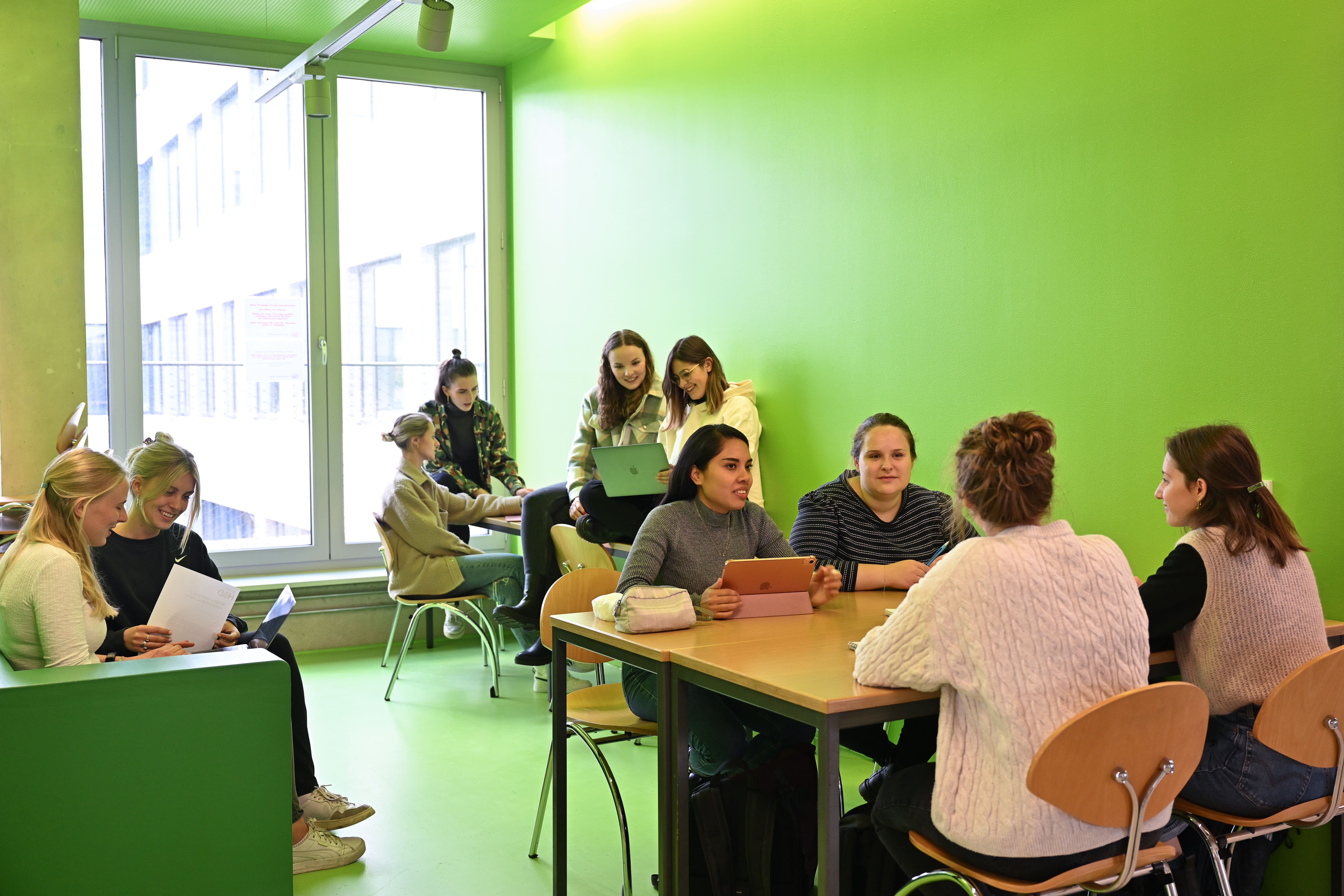 Studenten des Fachbereichs Sozial- und Kulturwissenschaften sitzen in einem grünen Raum und arbeiten in kleinen Gruppen mit Laptops, Tablets und Unterlagen zusammen.