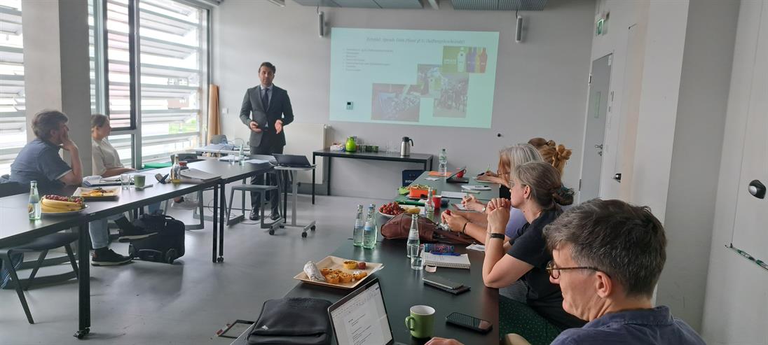 Personen sitzen an einer U-förmigen Tischformation und schauen den Sprecher in der Mitte an.