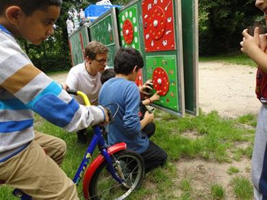 Auf diesem Bild sieht man einen Erwachsenen, der vor einer Tafel kniet, die mit roten und grünen Scheiben versehen ist. Neben ihm knien zwei kleine Jungen und zeigen darauf. Links hinter ihnen befindet sich ein Junge auf einem Fahrrad, der ihnen zu sieht. Rechts im Bild steht ein Mädchen, das ebenfalls zuschaut. 