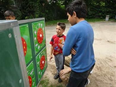 Auf diesem Bild sieht man im Mittelpunkt eine grüne Tafel mit roten Scheiben. Davor stehen zwei Jungen, welche diese betrachten. Hinter der Tafle steht noch ein Junge, von dem man nur den Kopf sieht. 