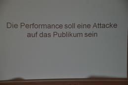 Zu sehen ist eine Powerpointfolie. Geschrieben ist: Die Performance soll eine Attacke auf das Publikum sein.