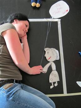 Zu sehen ist eine junge Frau die auf der Seite liegt und einer weiblichen Puppe aus Papier zugewandt ist. Symbolisch streckt die junge Frau ihre Hand in Richtung der Hand der Papierpuppe.