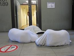 Zu sehen ist ein Flur des alten FH-Gebäudes. Auf dem Boden liegen drei Personen, dessen Körper vollständig von einem weißen Tuch umhüllt werden. 