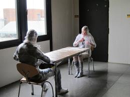 Zu sehen sind zwei Personen die sich an einem Tisch gegenüber sitzen. Beide sind bedeckt mit einer durchsichtigen Folie. 