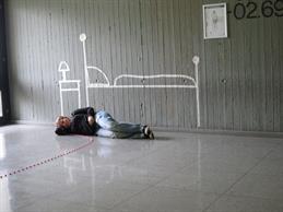 Zu sehen ist eine Person die in einem Flur auf dem Boden liegt und schläft. Auf der dahinter liegenden Wand ist mit Kreide ein Bett und ein Nachttisch aufgemalt. 