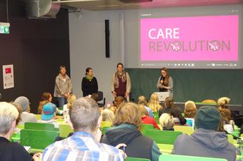 Gefüllter Vorlesungsraum mit grünen Stühlen. Ein Beamer projeziert das pinke Logo mit der Aufschrift "Care Revolution". Vorne stehen 4 Personen die Vortragen; der Rest sitzt auf den Stühlen.