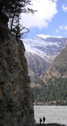 Zwei Wanderer passieren ein Tal zwischen schneebedeckten und bewälderten Bergen.