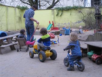 Auf dem Bild sind drei Kinder auf Spielzeugfahrzeugen und eine erwachsene Person von hinten zu sehen. Im Hintergrund befindet sich ein Spielplatz mit Sandkasten und weiteren Spielgeräten. Links stehen Kinderbänke. Das Setting ist von einer Mauer umgeben.
