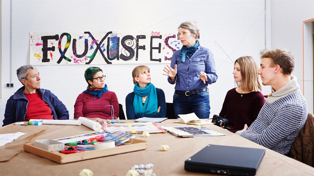 Students with a professor at a table with various utensils for artistic creation and before a long strip of paper on the wall with a painting of Fluxusfestspiele in various letters.