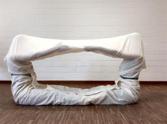 Zwei Personen gemeinsam eingehüllt in einen weißen schlauchförmigen Stoff. Sie beide bilden, in dem sie sich auf dem Boden gegenüber sitzen, ihre Arme nach vorn strecken eine geschlossene Form. 